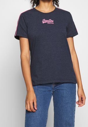 Kvinde iført navyblå SuperDry t-shirt med lyserød logo og striber på ærmerne, kombineret med blå jeans mod en ensfarvet baggrund.