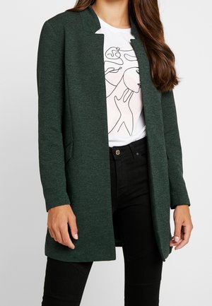 Femme portant un manteau vert foncé ouvert sur le devant, un t-shirt blanc avec un motif linéaire abstrait et un pantalon noir, debout devant un fond uni.