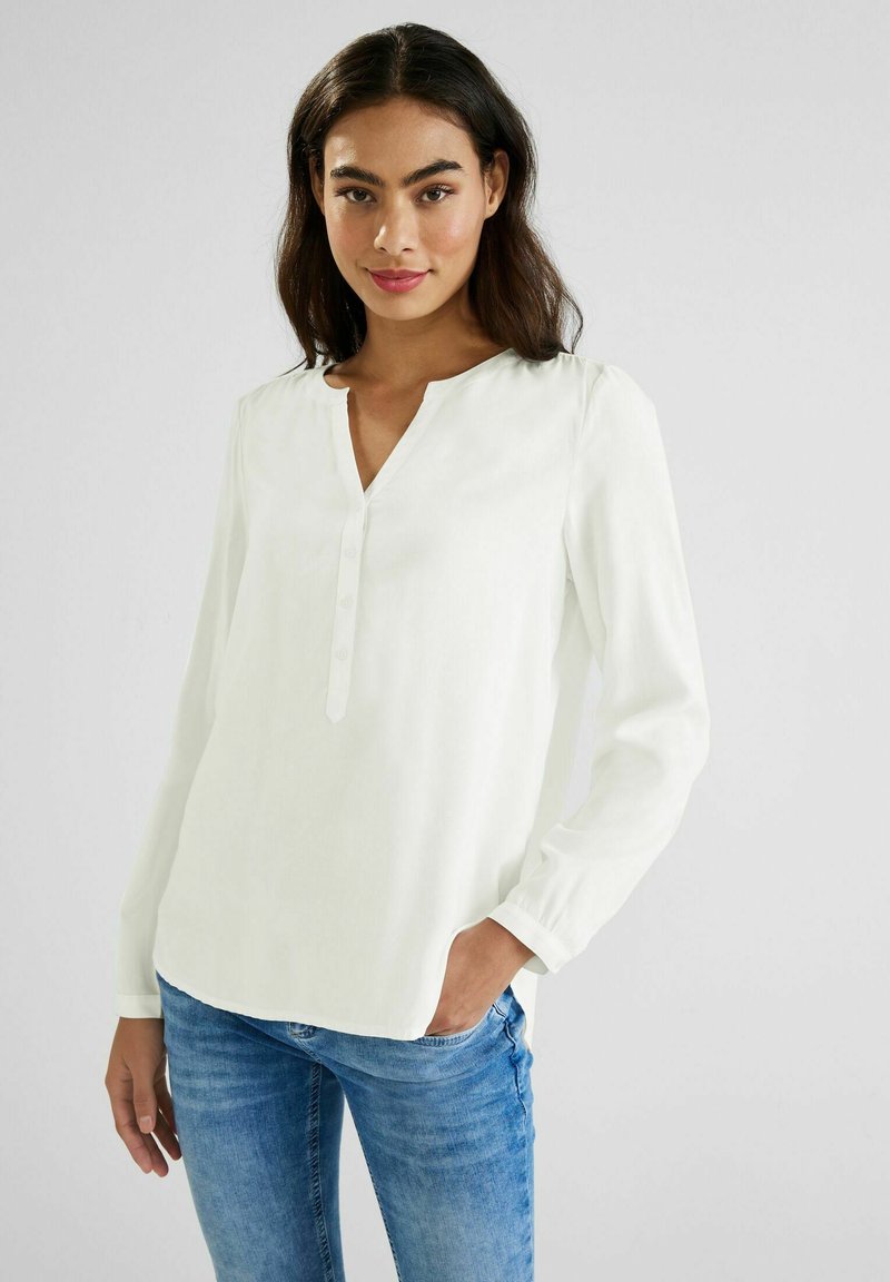 Blusa bianca a maniche lunghe con scollo a V, dettagli con bottoni e texture morbida. Indossata con jeans blu. Design semplice, tessuto leggero.