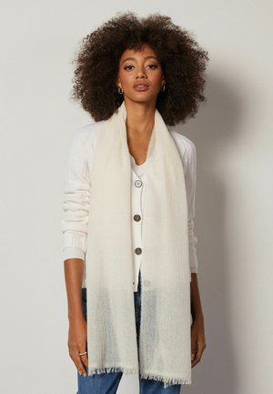 Elfenbensfarvet tørklæde lavet af blødt, tekstureret stof. Det har frynset kanter og falder elegant, båret over en lyse cardigan.