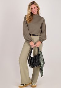Vrouw lacht, draagt een olijfgroene top met lange mouwen en beige wijde broek, houdt een zwarte handtas met studs vast met een groen gestreepte sjaal.