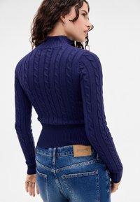 Knit navy blue sweater with a high collar, featuring cable patterns and ribbed cuffs. Worn with mid-rise blue jeans, showcasing a tag.