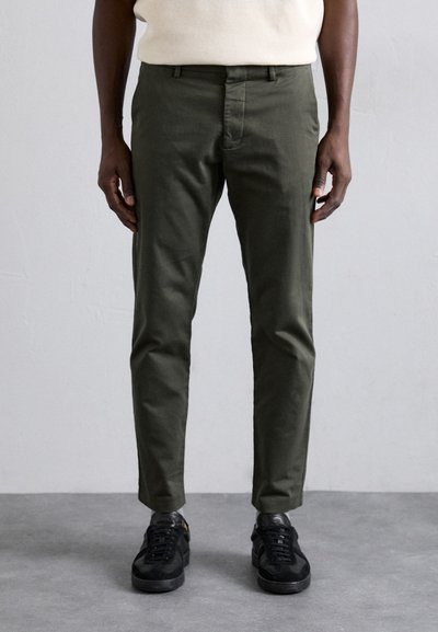 Pantalons en coton vert olive avec une coupe slim et un devant plat, dotés de passants de ceinture et de poches latérales, associés à des baskets noires.
