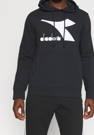 Person iført sort Diadora-hættetrøje med logo på brystet og matchende sorte bukser med hvide striber ned langs siden.