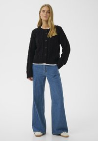 Fekete kabelmintás kardigán kerek nyakkal, gombos záródással és texturált mintázattal, széles lábú farmernadrággal és világos színű cipővel párosítva.