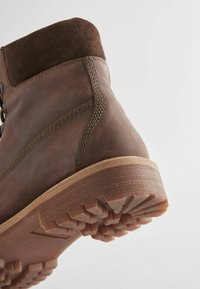 Bota de cuero marrón con un cuello de ante, suela resistente y un patrón de textura en la suela. Cuenta con costuras a lo largo del costado y un diseño robusto.