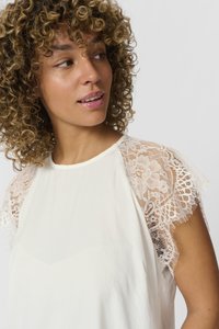 Top bianco con maniche in pizzo. Il pizzo presenta motivi floreali e un bordo sfrangiato, mentre il corpo è liscio e leggero.