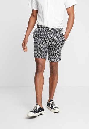 Mężczyzna ubrany w białą, krótkorękawową koszulę na guziki, czarno-białe szorty w wzory oraz czarne niskie trampki z białymi sznurowadłami i podeszwami.