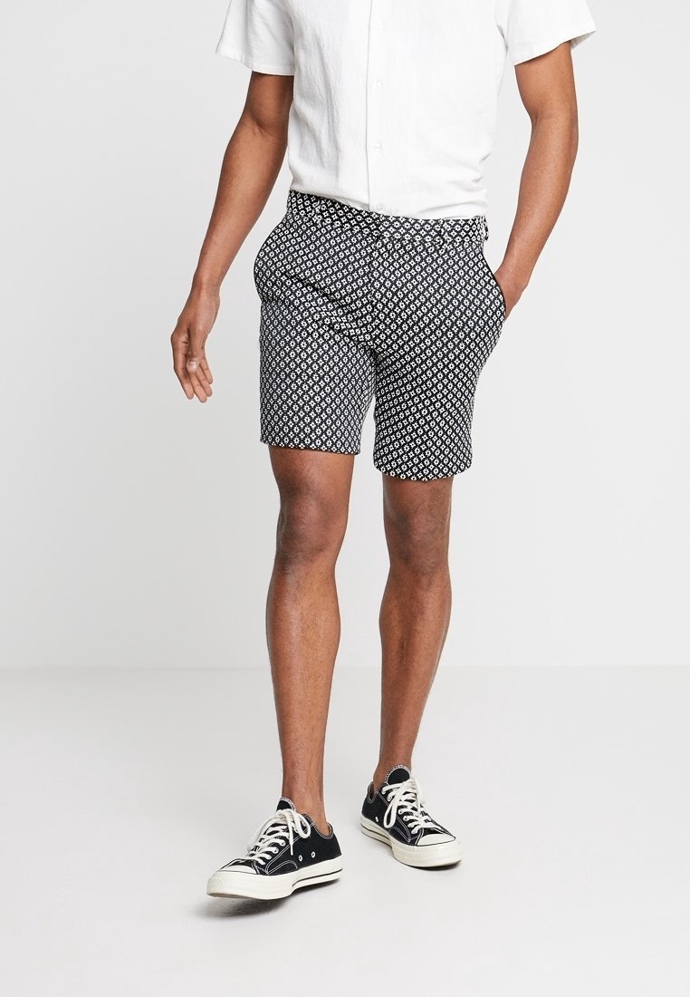 Homme portant une chemise blanche à manches courtes boutonnée, un short à motifs noir et blanc, et des baskets basses noires avec des lacets et semelles blanches.