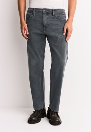 Person iført mørkegrå jeans med lige pasform og sorte lædersko, stående mod en ensartet lys baggrund.