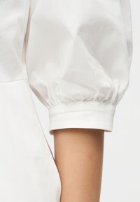 Λευκό μανίκι με φουσκωτό μανσέτα, φορεμένο σε γυμνό χέρι, που δείχνει την υφή του υφάσματος και τη λεπτομέρεια του μανικιού.