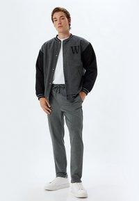 Giacca varsity grigia con maniche nere, lettera "W" sul petto, abbinata a pantaloni della tuta grigi e sneakers bianche. Tessuto liscio e vestibilità rilassata.