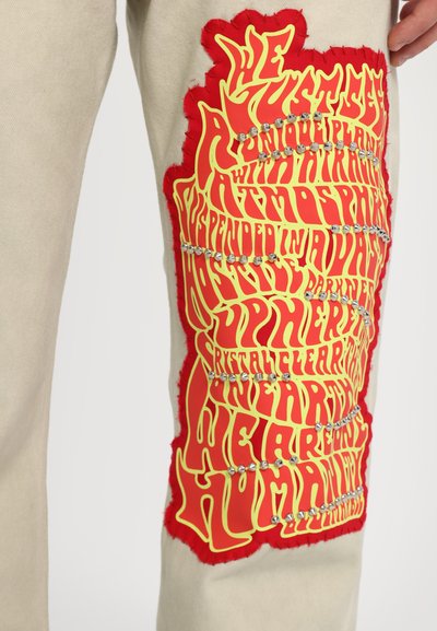 Beige trousers featuring a textured red patch with yellow graphic text. Adorned with small metallic studs for added detail.
