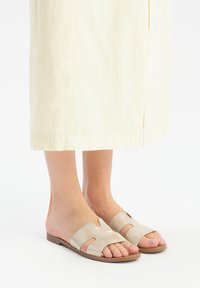 Beige színű, kivágott dizájnnal rendelkező lapos talpú szandálok. A modell világos sárga len szoknyával viseli őket.