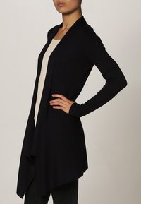 Marinblå ribbad cardigan med öppen frontdesign, långa ärmar och asymmetrisk nederkant, som visar en texturerad yta och minimal detaljering.