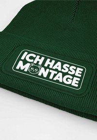 Green knitted beanie with a textured pattern, featuring a rectangular white label. The label includes black text and a cat graphic.