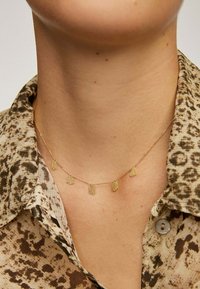 Collar de cadena dorada con cinco charms con textura, que se lleva contra un escote que presenta un cuello de camisa con estampado de animal en tonos marrones.