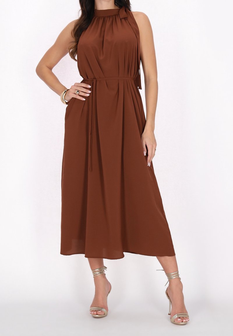 Femme portant une robe midi marron sans manches avec une ceinture nouée à la taille et des sandales dorées à talons hauts, posant avec une main sur la hanche.