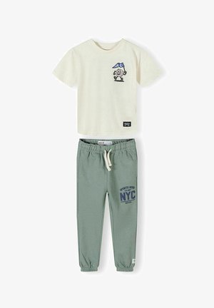 T-shirt crème à manches courtes avec un dessin animé tenant un drapeau de NYC et un pantalon de jogging vert avec cordon de serrage et impression sportive NYC sur la jambe gauche.