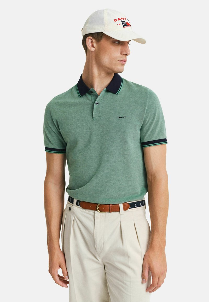 Camisa polo verde con un cuello azul marino y acentos a rayas, que presenta un logotipo en el pecho. Combinada con pantalones de pinzas de color claro.