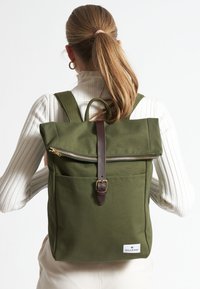 Mochila de lona verde oliva con diseño de parte superior enrollable, correa y hebilla de cuero marrón, bolsillo con cremallera y bolsillo lateral.