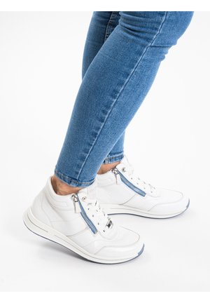 Persoon draagt blauwe skinny jeans en witte hoge sneakers met zijritsen, staand op een witte achtergrond.