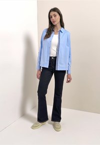 Camisa azul claro con botones y detalle de rayas, camiseta blanca sin mangas, vaqueros oscuros acampanados y zapatillas verde claro. Tejido suave, ajuste holgado.