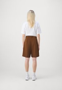Person med långt blont hår som bär en vit t-shirt, bruna knälånga shorts, vita strumpor och vita sneakers, stående vänd bortåt mot en enfärgad bakgrund.
