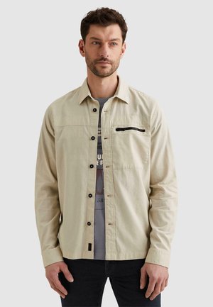 Muž nosiaci béžovú košeľu na gombíky s čiernym zipsovým vreckom cez sivé tričko s grafickým motívom a tmavé nohavice, stojaci pred jednoduchým pozadím.