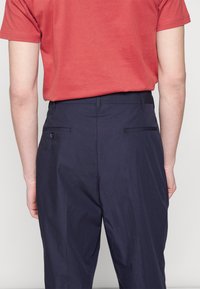 Mörkblå byxor i lätt tyg med rak passform, med två bakfickor och knapplåsning, iklädda med en röd t-shirt.