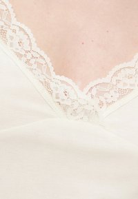 Gros plan sur un haut en tissu crème avec une bordure en dentelle florale complexe le long d'un décolleté en V sur une peau claire.