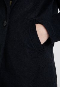 Dunkelblauer Mantel aus einem strukturierten, weichen Stoff, mit einem Knopfverschluss und einer Seitentasche. Nahaufnahme, die das Material und die Taschendetails hervorhebt.