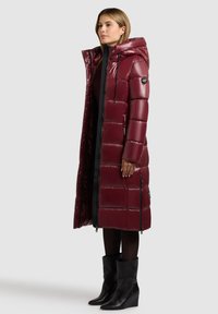 Burgundská dlouhá péřová bunda s lesklým povrchem, prošitým vzorem, kapsami na zip a odnímatelnou kapucí. Noshena přes černý outfit a boty.