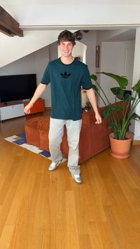 T-shirt de mangas curtas azul-petróleo com logótipo preto, jeans cinza-claro e sapatilhas prateadas. Em pé num chão de madeira, com um sofá castanho e uma planta próximos.