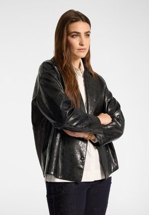 Donna con lunghi capelli castani che indossa una giacca di pelle nera, una camicia bianca, pantaloni scuri e un braccialetto d'oro, in piedi con le braccia incrociate.