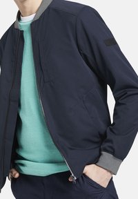 Námořnická bomber bunda s šedým ribovaným límcem a manžetami, která má přední zip a boční kapsu, vrstvená přes strukturovaný svetr v mátově zelené barvě.