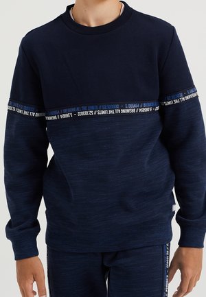 Junge trägt einen marineblauen Pullover und passende Hose mit Textbändern mit der Aufschrift "BREAKING ALL THE LIMITS" über Brust, Ärmeln und Hosenbeinen.