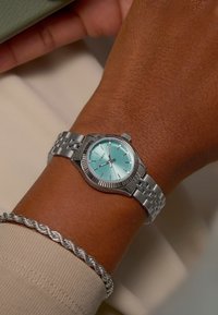 Silver wristwatch with a mint-green dial, metallic indices, and fluted bezel. Bracelet features interlocking links. Worn on a wrist with a silver bracelet.