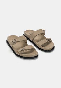 Suede slip-on sandals in beige with two adjustable straps and metallic buckles, featuring a black rubber sole. Textured surface.