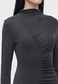 Femme portant un haut gris foncé à manches longues avec un design froncé asymétrique et un col montant, vue des épaules jusqu'au milieu du torse.