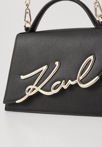 Czarna skórzana torba o wyrazistym kształcie, z metalicznym złotym akcentem w postaci logo oraz łańcuszkiem z zapięciem.