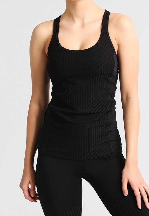 Femme portant un débardeur de sport noir sans manches et un legging noir, debout, les bras détendus, devant un fond gris clair uni.