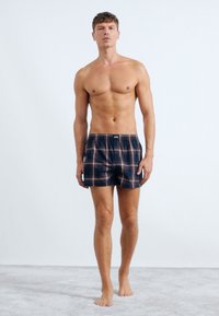 Boxer da uomo a quadri in blu navy e bordeaux, con vita elastica e vestibilità comoda. Modello in posa in piedi su una superficie grigia chiara.