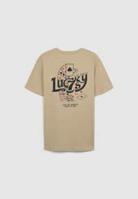 T-shirt in cotone beige con una stampa grafica di carte da gioco e il testo "Lucky 7's Las Vegas" in colori nero e rosso. Maniche corte, vestibilità rilassata.