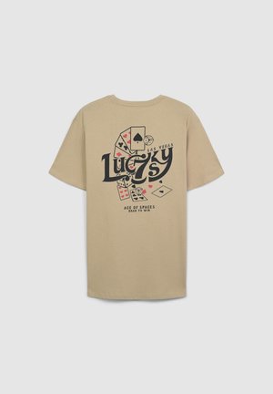 T-shirt in cotone beige con una stampa grafica di carte da gioco e il testo "Lucky 7's Las Vegas" in colori nero e rosso. Maniche corte, vestibilità rilassata.
