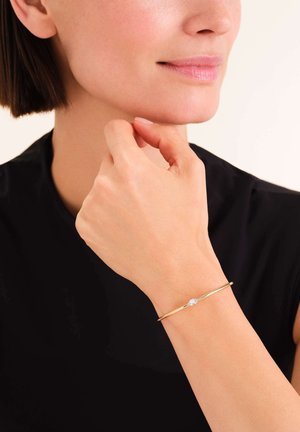 Pulsera de oro con un solo acento de piedra preciosa transparente. Acabado de metal suave y diseño minimalista, llevada en una muñeca.