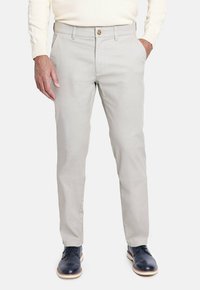 Hombre con pantalones slim-fit beige claro, camisa blanca de manga larga y zapatos de vestir negros, con las manos en los bolsillos, de pie sobre un fondo blanco.