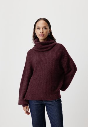 Burgunderfarget strikkegenser med høy rullet hals og vide ermer. Har et teksturert mønster og en avslappet passform. Kombinert med mørke jeans.