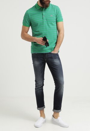 Homme portant un polo rayé vert, jean foncé retroussé, chaussures blanches sans lacets, tenant des lunettes de soleil noires, debout devant un fond uni.