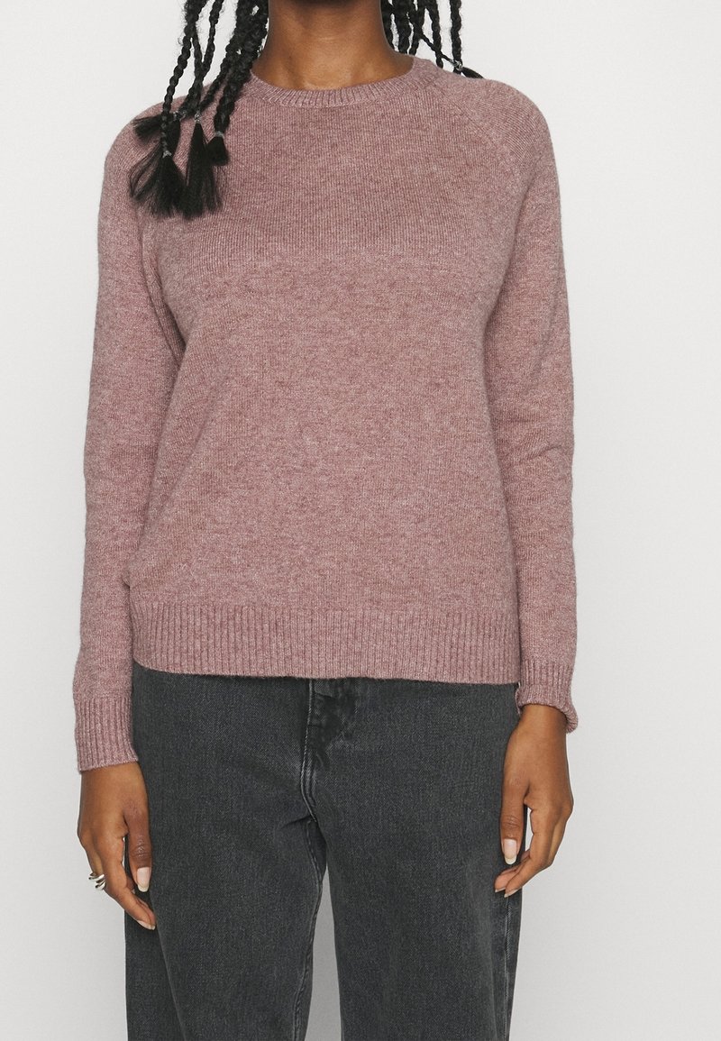 Personne portant un pull à manches longues mauve en tricot avec un col rond et un jean noir taille haute, montrant le buste et les bras avec des cheveux tressés qui tombent.